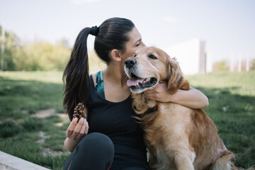 Woman cuddling her dog in city park. Pretty woman cuddling and kissing dog while sitting on lawn in park and holding pinecone.