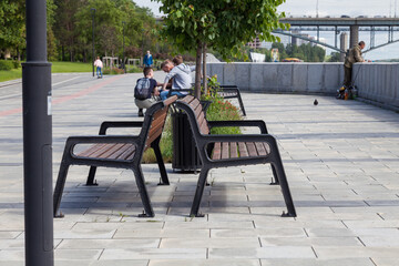 Fototapeta na wymiar City park with a promenade covered with paving slabs and wooden benches where people relax and against the background of a car bridge and a fisherman and a child riding on skateboard on bicycle path.