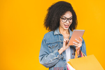 Shopping Concept - Portrait young beautiful african american woman smiling and joyful with colorful shopping bags and tablet computer isolated over yellow Background. Copy Space.