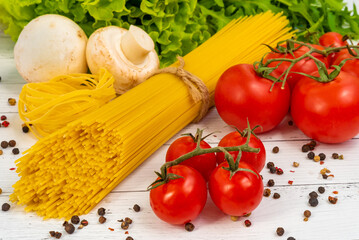 On the table are spread out spaghetti, tomatoes, herbs, peppers, garlic, and mushrooms.