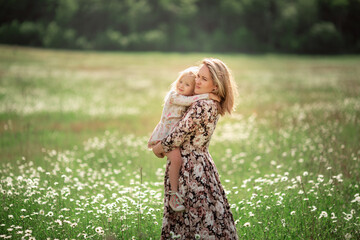 Beautiful young mother with her daughter on a field with daisies sunset sun, life style, concept of motherhood, walk in the park or in nature.