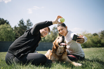 Women with phone and ball playing with dog outdoors. Beautiful women taking selfie with phone while sitting on lawn and playing with dog.