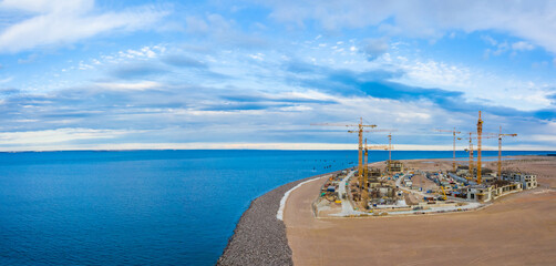 Fototapeta premium Panorama of a building near the sea. Many tower cranes on seashore. Construction is underway on artificially created island. Aerial survey of a construction site. Erection of buildings on the beach
