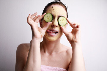 Happy young woman with a face mask on holding a cucumber slices in front of eyes. Beauty and...