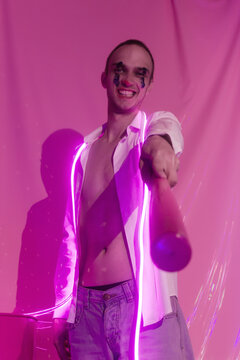 Portrait of Young Handsome emotional man in an unbuttoned white shirt with pink bat. Male in the image of a crazy clown like a joker. Neon pink room. Reflect of neon. Selective focus.