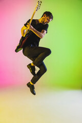 In jump. Young caucasian musician playing bass guitar on gradient studio background in neon. Concept of music, hobby, festival. Colorful portrait of modern artist. Inspired, impressive improvising.
