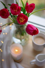 Still life coffee at home, a cozy coffee, tulips in a vase. Coffee on the windowsill, comfort at home. Milk jug  mug, candlestick, envelope and postcard on wooden desk. Spring content