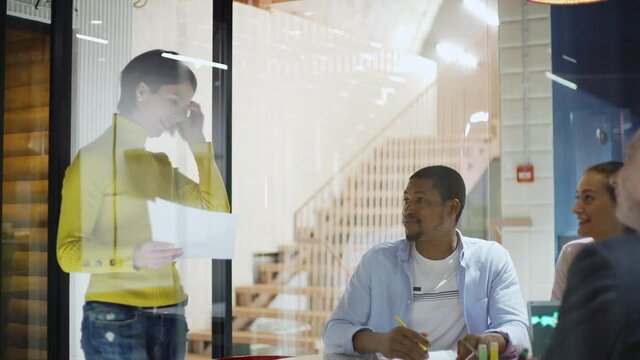 Tracking medium shot of middle aged businesswoman entering office behind glass wall and presenting printouts with her ideas to colleagues sitting at table