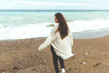 Fototapeta na wymiar A beautiful young girl walks along the seashore, a storm, hair fly apart, a gray cardigan, a sports figure, in sneakers, enjoys, autumn or winter