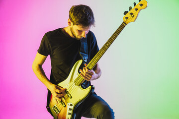 Close up young and joyful caucasian musician playing bass guitar on gradient studio background in neon light. Concept of music, hobby, festival. Colorful portrait of modern artist. Inspired - obrazy, fototapety, plakaty