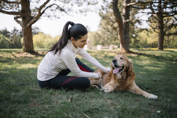 Woman in sportswear petting her retriever dog. Pretty girl and dog lying on ground in park near trees during sunny day.