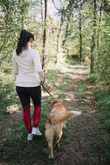 Back of girl and dog walking on forest road. Woman dressed in sport outfit walking her dog in nature during sunny day.
