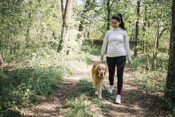 Girl wearing sport outfit holding her dog on leash. Sport woman having a walk with her dog in nature during sunny day.