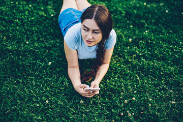 .Attractive hipster girl with braid resting on grass while checking mail on smartphone