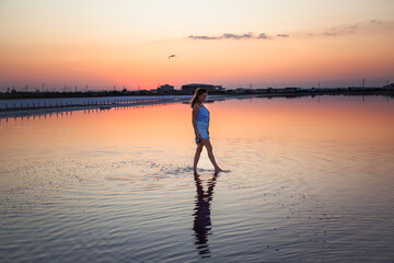 Fototapeta premium Rear view of the woman back silhouette look to the purple sunrise in water on summer. Woman walking on the shore of the salt beach at pink lake. vacation lifestyle near ocean