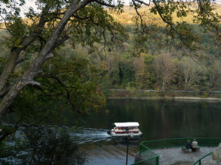 A couple sitting on Olt River's bank, Romania, in the evening, tourists boat cruising pass by.