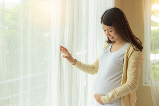 Happy pregnant woman touching and looking fetus at belly with smile stand near window at home. prenatal, pregnancy, motherhood, expect concept. Young pregnance girl care and love her baby copy space