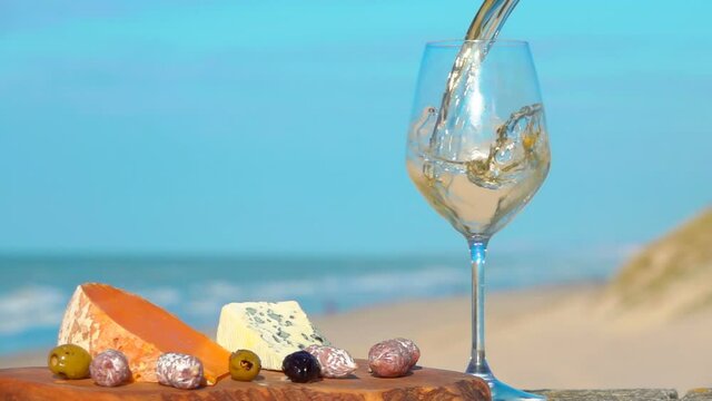 White wine is poured in the glass next to the various sorts of cheese on the background of the sea. Romantic picnic by the sea 