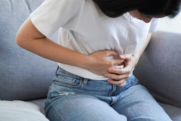 Fototapeta na wymiar Young Asian woman suffering from strong abdominal pain stomachache or period comes while sitting on sofa at home