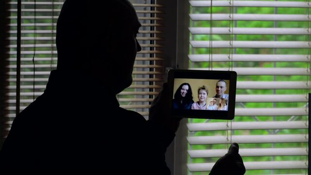 A bald man is facing the light at a window with blinds, she is talking on a video conference on a tablet with a group of different people: a woman mom, a man dad and a little girl, a young couple.