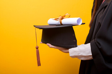 Close up of woman hand in graduation gown is holding graduation cap and certificate on yellow...
