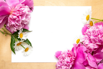 White paper card on rustic wooden background with pink peonies and white chamomile. Flowers. Workspace. Blank for greetings