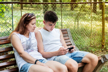 Teenager sits on a park bench and plays games on the phone, mom sits next to him and also looks at the phone, she wants to talk to her son and he is busy