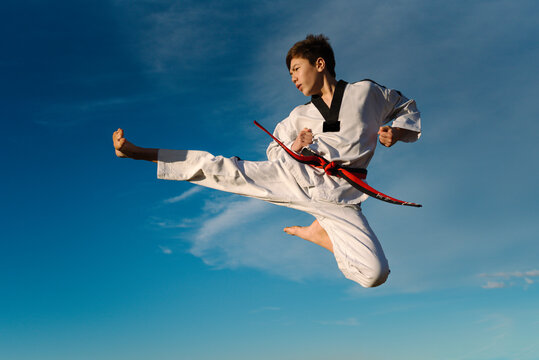Karate boy kicks in the air and flies over the blue background of the sky. Practicing Taekwondo in nature