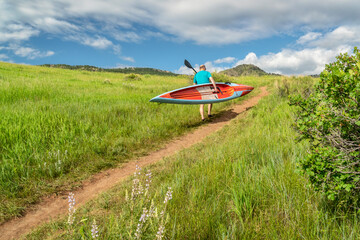 senior male paddler is carrying a long racing stand up paddleboard (SUP) uphill on a trail in early...