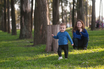 A young mother smiles at her one year and a haly years old son as he walks through a park.