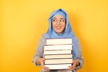 Young beautiful muslim woman wearing hijab and holding books on her arms against yellow background looking aside being happy and glad to pass her exams. Confidence, college and religion concept. 