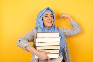 Young beautiful muslim proud woman wearing hijab and holding books on her arms against yellow...