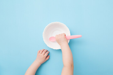 Baby hand putting pink plastic spoon in empty bowl and waiting food on light blue table background....