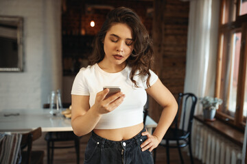 Indoor shot of displeased plus size teenage girl with wavy hair holding cell phone, having...