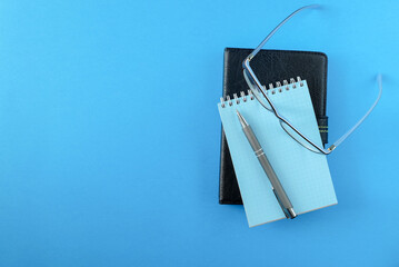 A daily planner or e-book in a black leather cover, a notepad, glasses and a pen on the blue surface. Horizontal orientation. View from above, copy space.