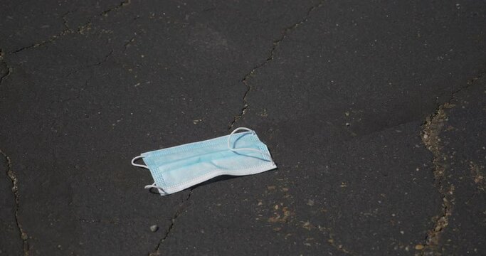 A closeup view of a used surgical mask being carelessly dropped in a store's parking lot. Shot at 48fps slow motion.  	