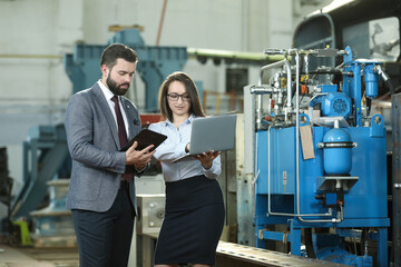 Portrait of a solid businessman with his secretary holding laptop and tablet, talking about factory financial report. Industrial workers.