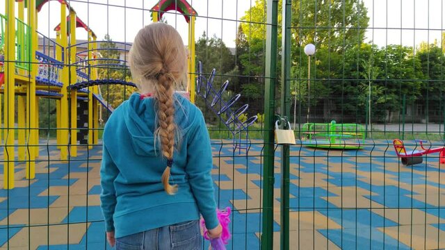 Sad lonely child near the closed playground because of the quarantine.A girl with long blonde braided hair staying alone near empty fenced showground without children.COVID-19 lockdown social distance