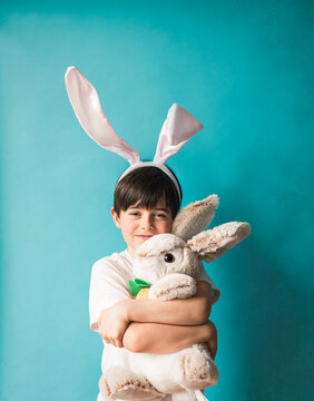 Boy wearing bunny ears hugging toy rabbit against blue background.