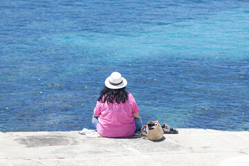 Fototapeta na wymiar Summer wanderlust concept off a woman sitting looking at a seascape in the Mediterranean Sea is wearing a hat and a pink T-shirt.
