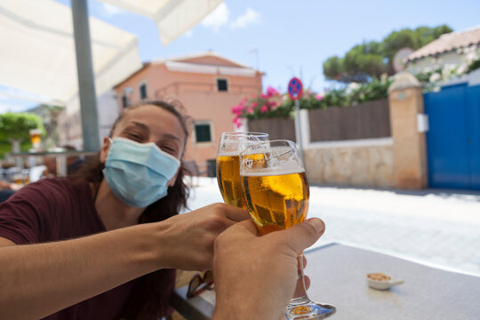 New Normality In Bars. A Couple Of Friends Toasting And Drinking Beer At A Bar. The Woman Is Wearing A Surgical Mask And They Are Celeb.rating The End Of Confinement