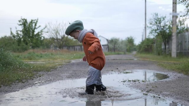 joyful childhood, healthy child in hat and rubber boots has fun playing and jumping into puddle on road after rain in countryside