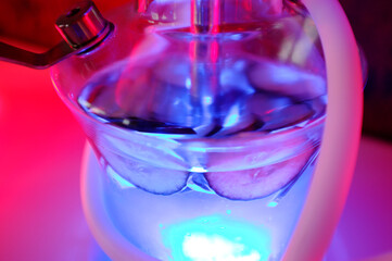 Colorful glass hookah water pipes with hoses on table, traditional shisha used for smoking in lounge bar. Various vibrant LED lights olluminating modern hookah setup.