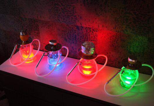 Colorful glass hookah water pipes with hoses on table, traditional shisha used for smoking in lounge bar. Various vibrant LED lights olluminating modern hookah setup.