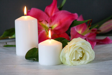 Fototapeta na wymiar Close up of two pillar burning candles and white rose on dark wooden table with pink lily flowers on the background. Memorial and remembrance concept.
