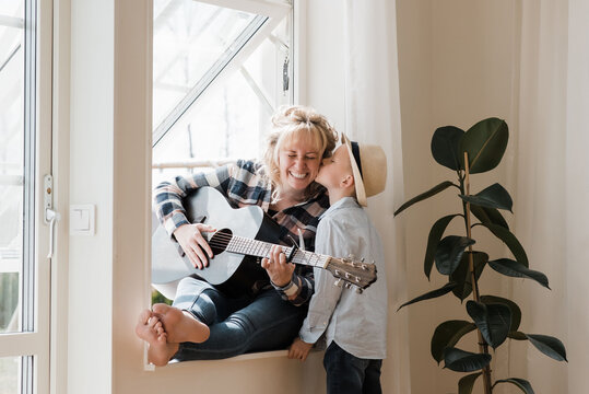 woman sat playing guitar at home whilst her son gives her a kiss