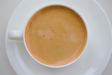 A white ceramic cup with a handle on a saucer and a coffee drink with a light brown pleasant foam. Close up, top view