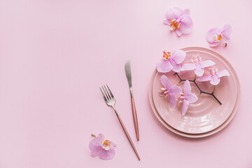 Flower and table settings overhead composition on light pink background. Pink ceramic plates, cutlery and perple orchid flowers. Holiday modern table decoration. Romantic date.