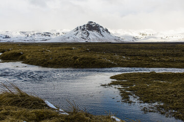 Glacial river flowing through volcanic black rock with mountains full of snow on the background