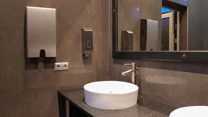 Chrome faucets with white round washbasins in a public toilet with a large mirror and gray walls,...